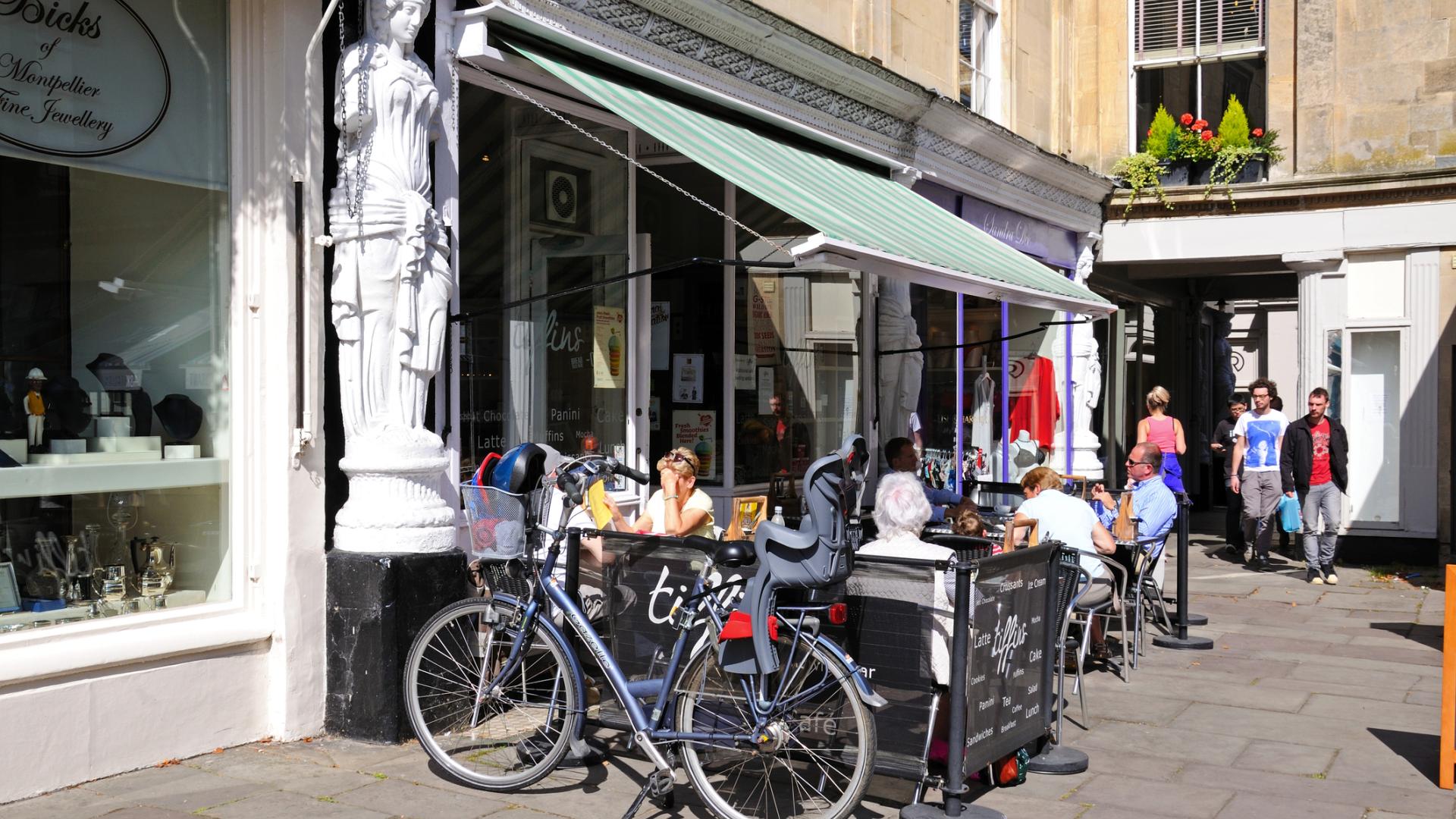 Street view of outdoor cafe tables with bike parked in foreground, people having coffee in sunshine