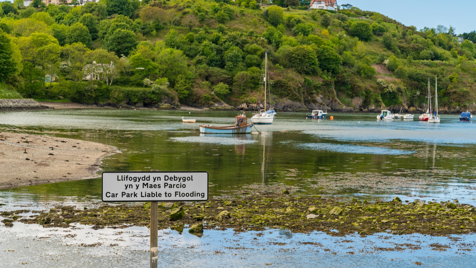 Sign in English and Welsh in front of flooded car park by river estuary, wooded slopes in background