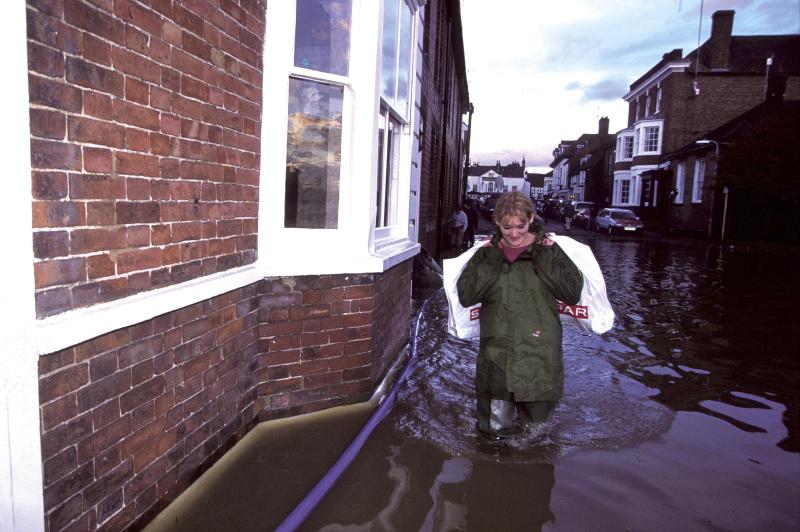 Woman wading in knee deep water past bay window in flooded street