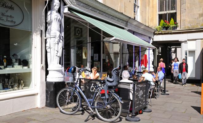 Street view of outdoor cafe tables with bike parked in foreground, people having coffee in sunshine