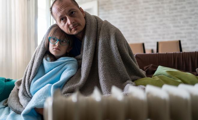 Cold father and daughter wrapped up in blankets on their sofa by a radiator